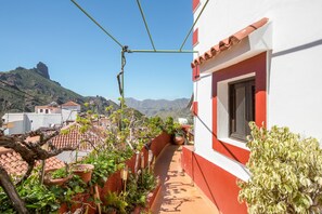 A narrow balcony with potted plants, a red and white building, and a mountainous backdrop.