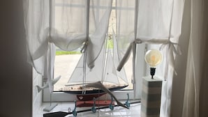 Et værelse med et vindue, let gardin, en modelsejlbåd og en lampe på vindueskarmen.