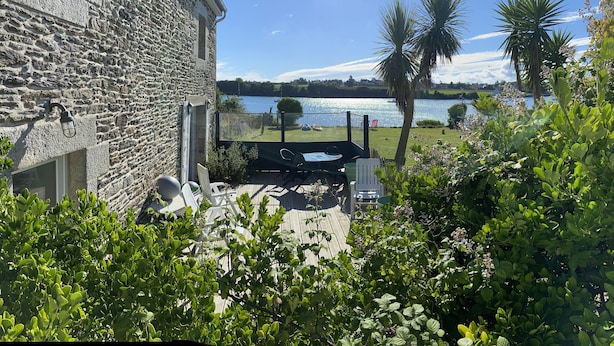 En bygning i sten med en patio med bord og stole, med udsigt over vand og græs.