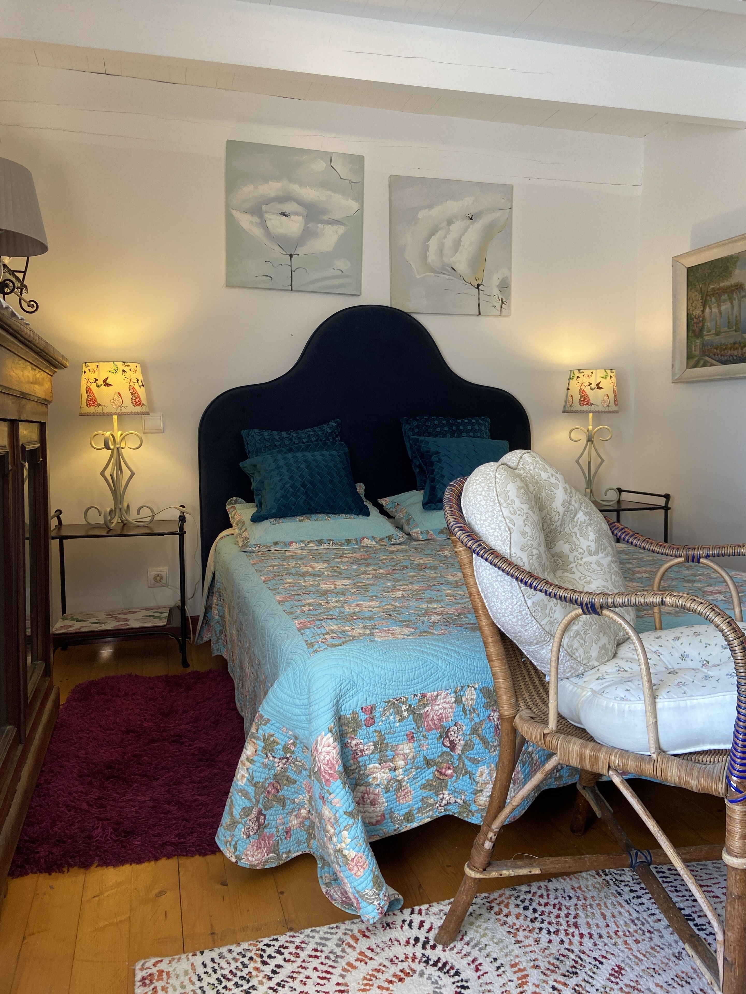 Ein Schlafzimmer mit einem großen Bett, zwei Nachttischen, einem Korbstuhl, einem roten Teppich und Wandbildern.