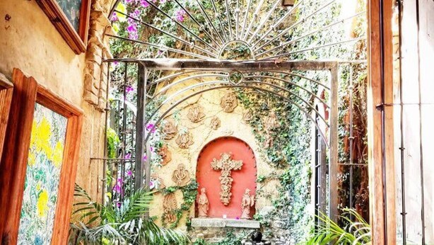 Un arco de entrada conduce a un patio con una fuente, rodeado de plantas en macetas y una puerta roja.