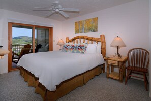 Quarto com uma cama grande, cabeceira de madeira, duas mesas de cabeceira com abajures, uma cadeira e vista para o exterior por uma porta deslizante.