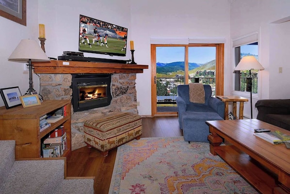 Uma sala de estar com lareira, televisão, sofá e vista para o exterior.