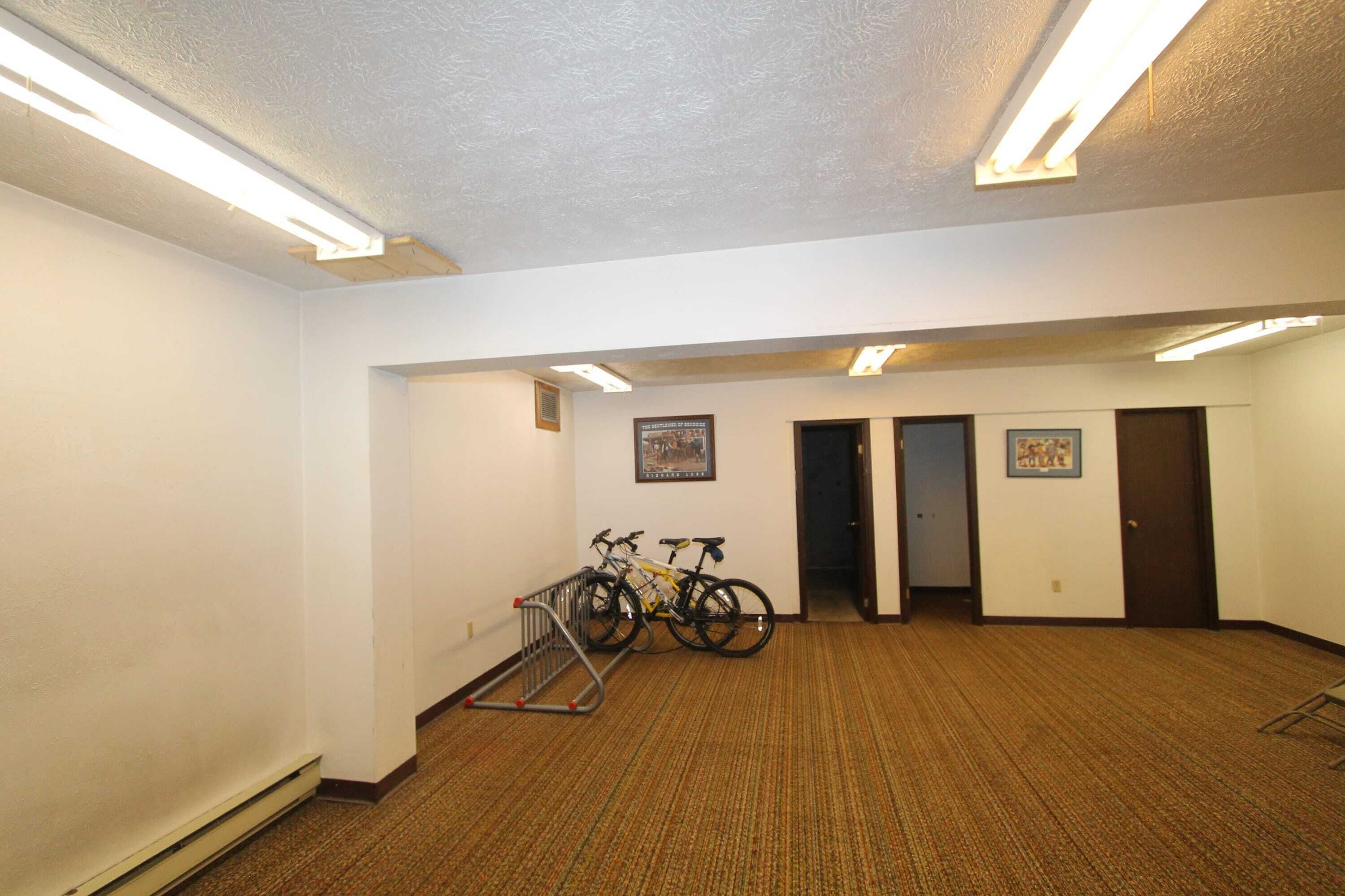 A room with carpeted flooring, fluorescent ceiling lights, two doors, and bicycles parked against the wall.
