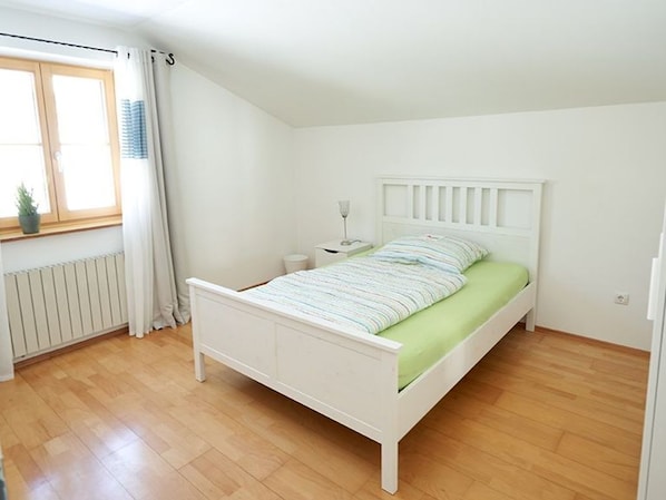 A bedroom with a white bed, a bedside table, a window with curtains, and a radiator.