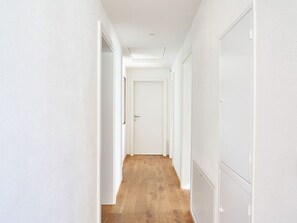 A narrow hallway with white walls and wooden flooring, leading to a closed door at the end.