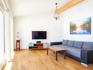 A living room with a wooden floor, a grey sofa, a TV mounted on the wall, and a painting of a landscape.