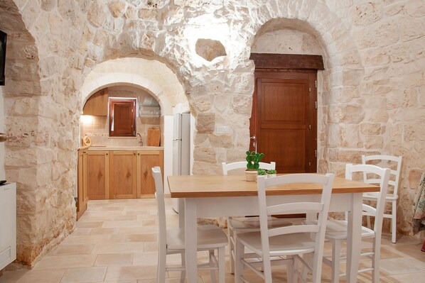A stone-walled room with a wooden table and chairs, a kitchen area in the background, and a large wooden door.
