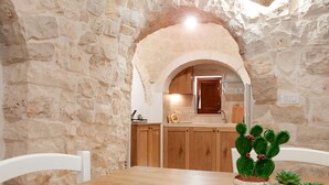 Una habitación con paredes de piedra, una mesa y sillas de madera, una zona de cocina con armarios de madera y una planta de cactus sobre la mesa.