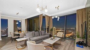 A modern living room with a grey sofa, white armchairs, and a coffee table. There is a dining area with a table and chairs, and large windows offering a city view. - FIRST CLASS | 3BR | Burj Khalifa & Fountain view (Downtown)