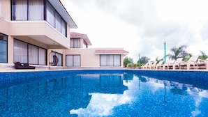Una piscina con agua azul cristalina, rodeada de sillas de descanso y un edificio con ventanas amplias y un balcón.