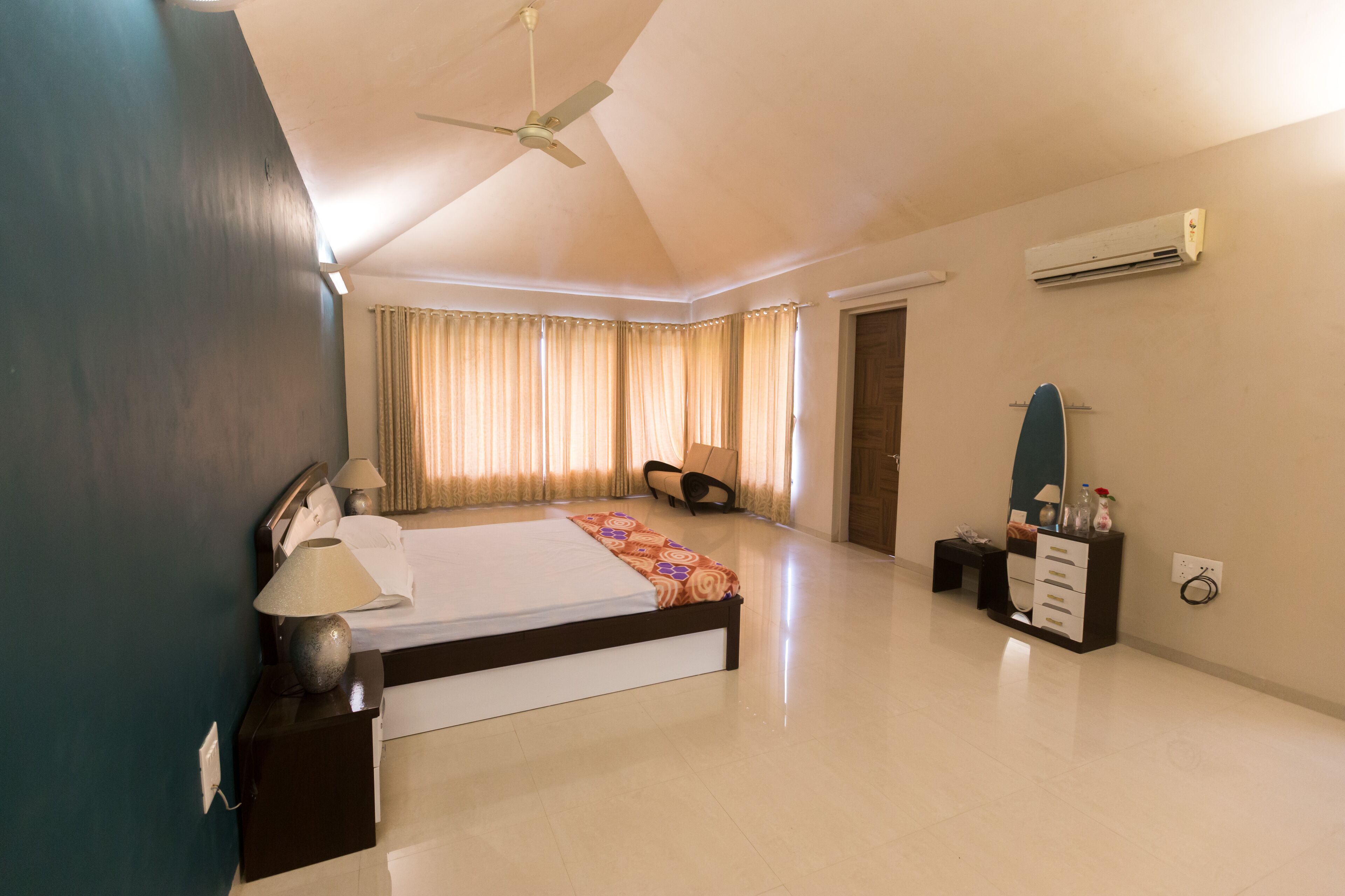 A spacious bedroom with a large bed, a ceiling fan, an air conditioning unit, a bedside table with a lamp, a dresser, and a chair.