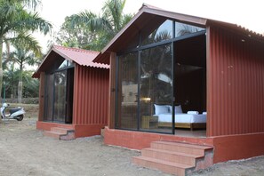 Two small, single-story cabins with large windows and a red roof, situated in a natural setting with palm trees.