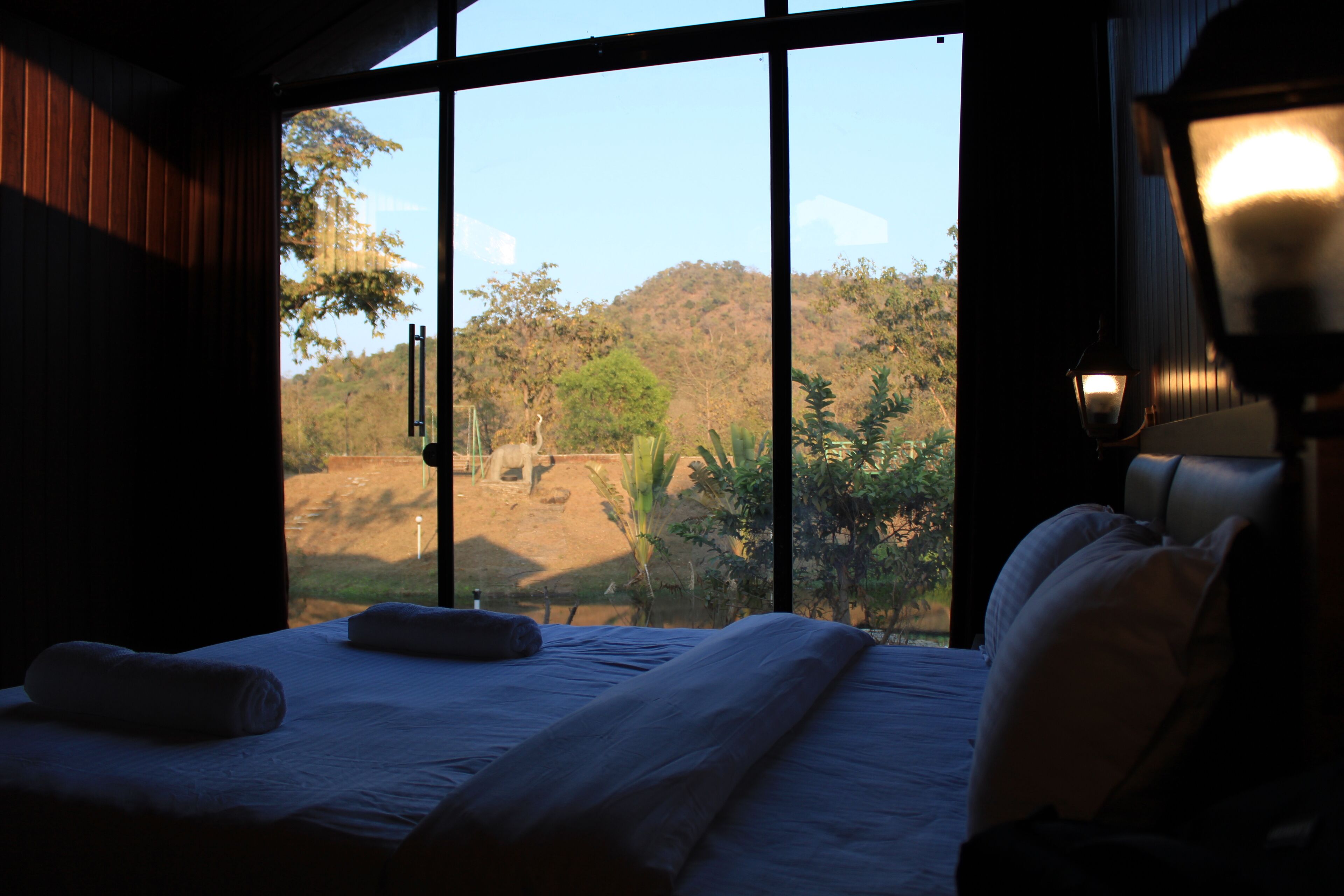 A bedroom with a large window, a bed with pillows, and a nightstand with a lamp.
