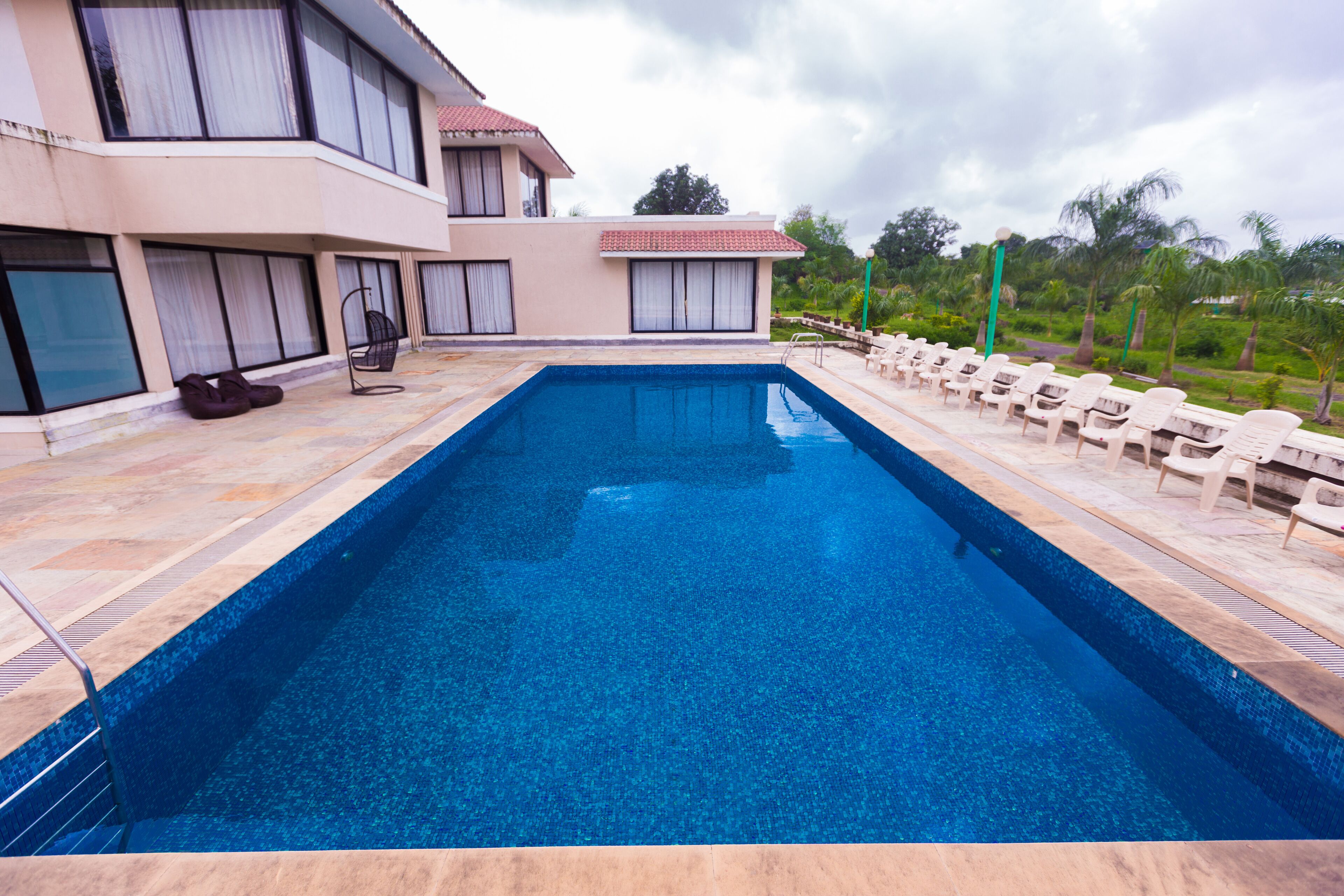 A rectangular swimming pool with a tiled edge, surrounded by lounge chairs and a building with large windows and a red-tiled roof.