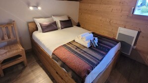 Ein hölzernes Bett mit weißer und brauner Bettwäsche, ein kleiner Holzstuhl und ein Fenster mit Blick auf Bäume.