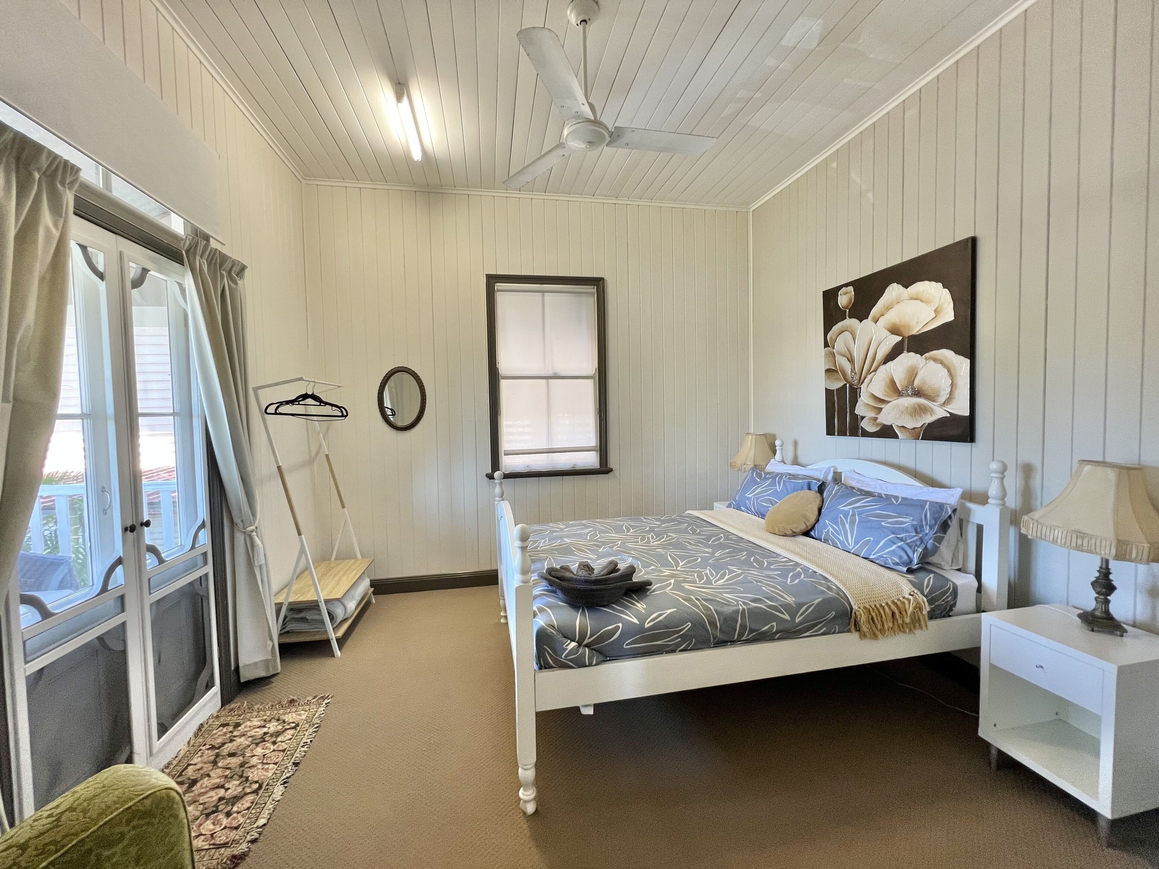 A bedroom with a bed, a nightstand, a hanging lamp, a window with curtains, a ceiling fan, and a painting on the wall.