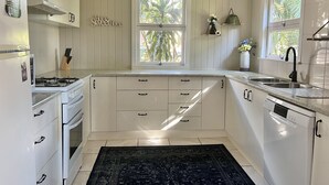 A kitchen with white cabinets, a gas stove, a dishwasher, and a window with a view of palm trees. - Childers charmer - with aircon, WIFI and modern luxuries (Childers)