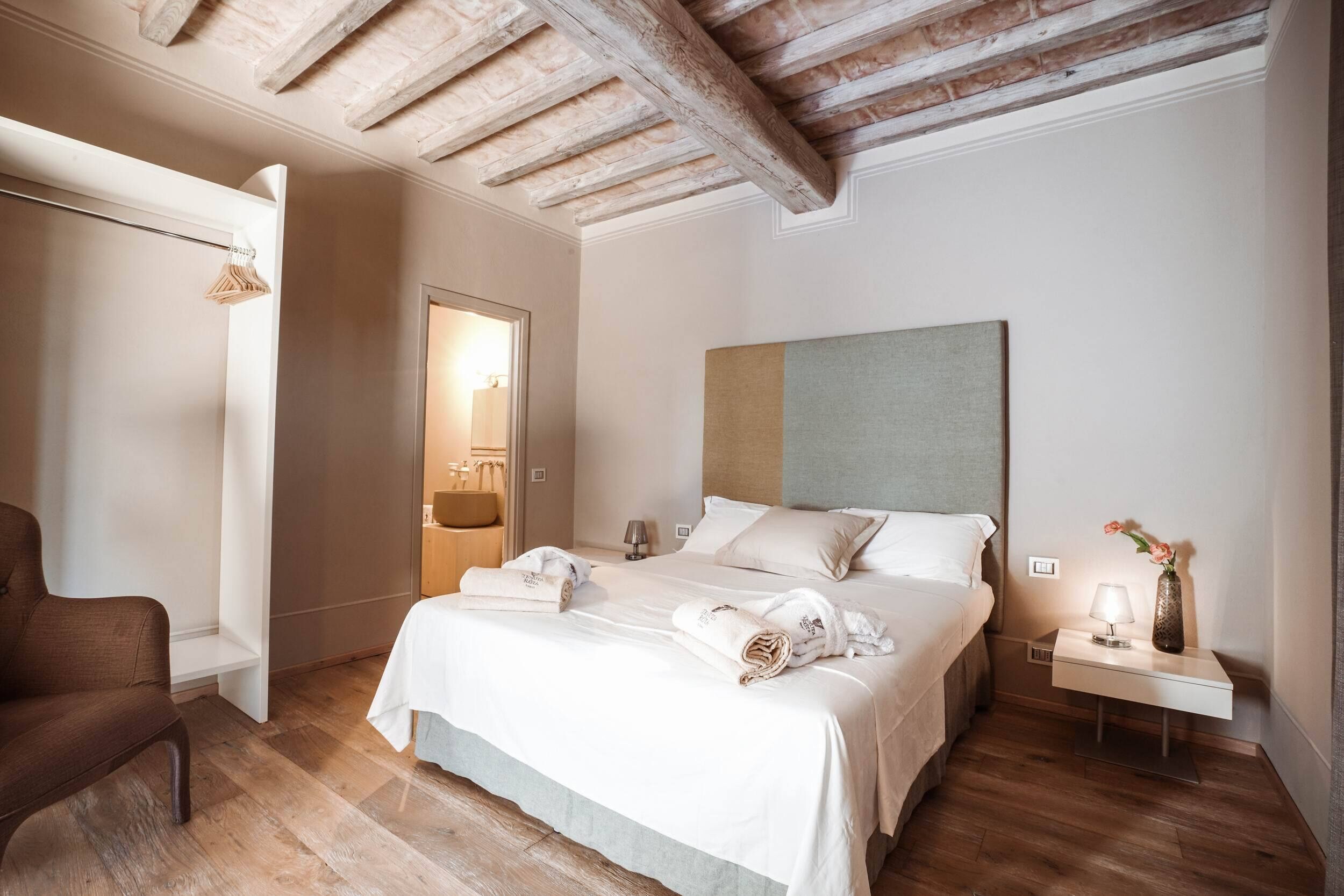 Une chambre d’hôtel avec un plafond en bois, un lit avec du linge de maison blanc, une table de chevet avec une lampe et un vase, et une chaise.