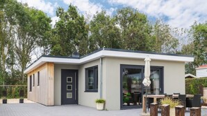 Een modern gelijkvloers huis met plat dak, een donkere voordeur en een grote glazen deur die leidt naar een patio met buitenmeubilair.