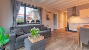 Een moderne woonkamer met een grijze bank, een houten bijzettafeltje met een bloevas en een groot raam met gordijnen.