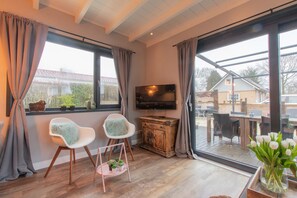 Un salon moderne avec un téléviseur, deux fauteuils blancs, un meuble en bois et une grande fenêtre avec des rideaux.