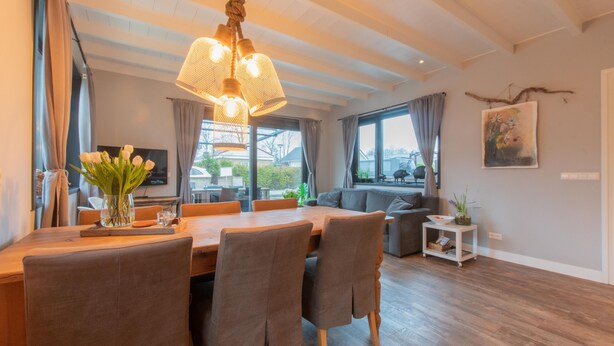 Een moderne woonkamer met een houten eettafel, zachte zithoek, een groot raam met gordijnen en een hanglamp.