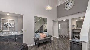 Een ruime woonkamer met een grijze bank, een grijze zithoek, en een keuken op de achtergrond.
