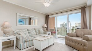 Un salon moderne avec un canapé à rayures, une table basse blanche et un canapé beige. On y trouve un ventilateur de plafond, une grande fenêtre avec des rideaux et une vue sur les bâtiments à l’extérieur.