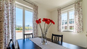 A dining table with a vase of red flowers, two chairs, and a view of a garden through a window with floral curtains. - Comfortable villa with private swimming pool (Marina di Ascea)