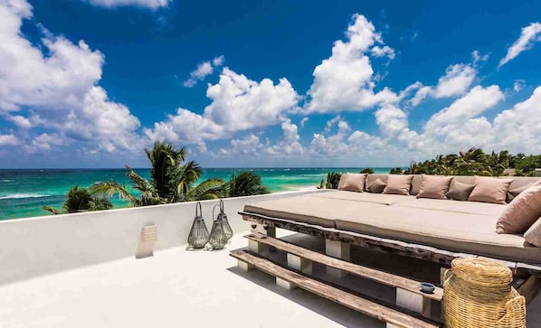 Relax in a serene oceanfront seating area with a sofa, pillows, and a wooden bench, surrounded by a tropical landscape.