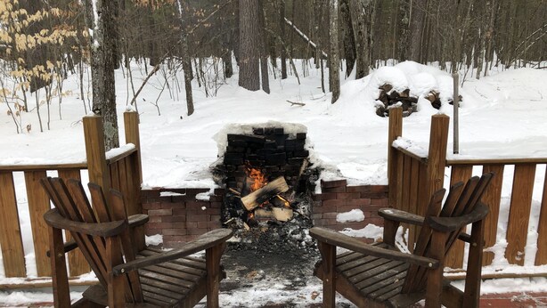A fireplace with two wooden chairs facing it, surrounded by a wooden deck and snow-covered ground.