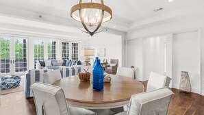 A spacious living room with a round wooden table, comfortable seating, and a large window with a view of greenery.