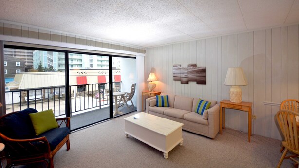 A living room with a sofa, a coffee table, a lamp, and a chair. There is a balcony with a view of buildings and a table outside. - One-bedroom and one and a half bath condo on ocean block! (Ocean City)