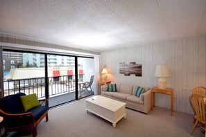 Ein Wohnzimmer mit einer Couch, einem Couchtisch, einer Lampe und einem Sessel. Ein Balkon bietet Blick auf Gebäude und einen Tisch im Freien.