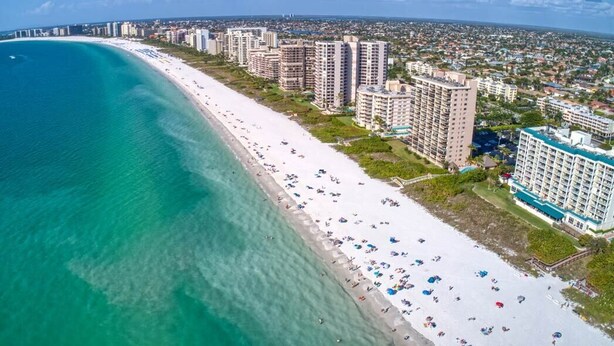 Vista aérea de una ciudad costera con una extensa playa de arena blanca, rascacielos y un mar azul cristalino.