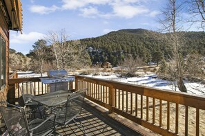 Un terrazzo in legno con mobili da esterno, un barbecue e vista su un paesaggio innevato con alberi e una collina.