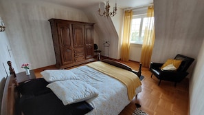 A bedroom with a large wooden wardrobe, a bed with a patterned bedspread, a black chair, and a window with curtains. - Charming property 3 km from the beach, with a covered swimming pool (Cléder)