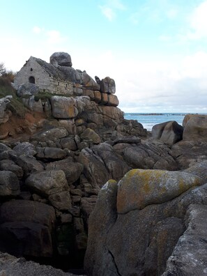 A stone structure with a small window, surrounded by large rocks and a body of water in the background. - Charming property 3 km from the beach, with a covered swimming pool (Cléder)