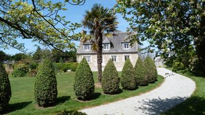 A stone house with a gravel driveway, surrounded by well-manicured hedges and trees.