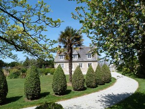 A stone house with a gravel driveway, surrounded by well-manicured hedges and trees.