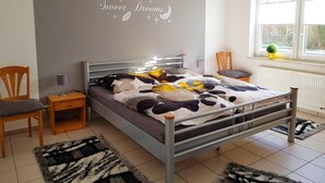 Une chambre à coucher comprenant un lit, deux chaises, une table de chevet, un mur orné d’une inscription décorative, une fenêtre donnant sur un paysage agréable et un radiateur.