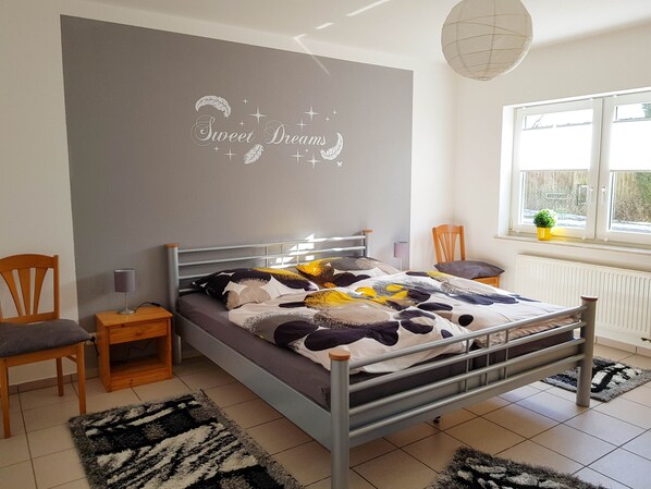 Un dormitorio con cama, dos sillas, una mesita de noche, una pared con una frase decorativa, una ventana con vistas y un radiador.