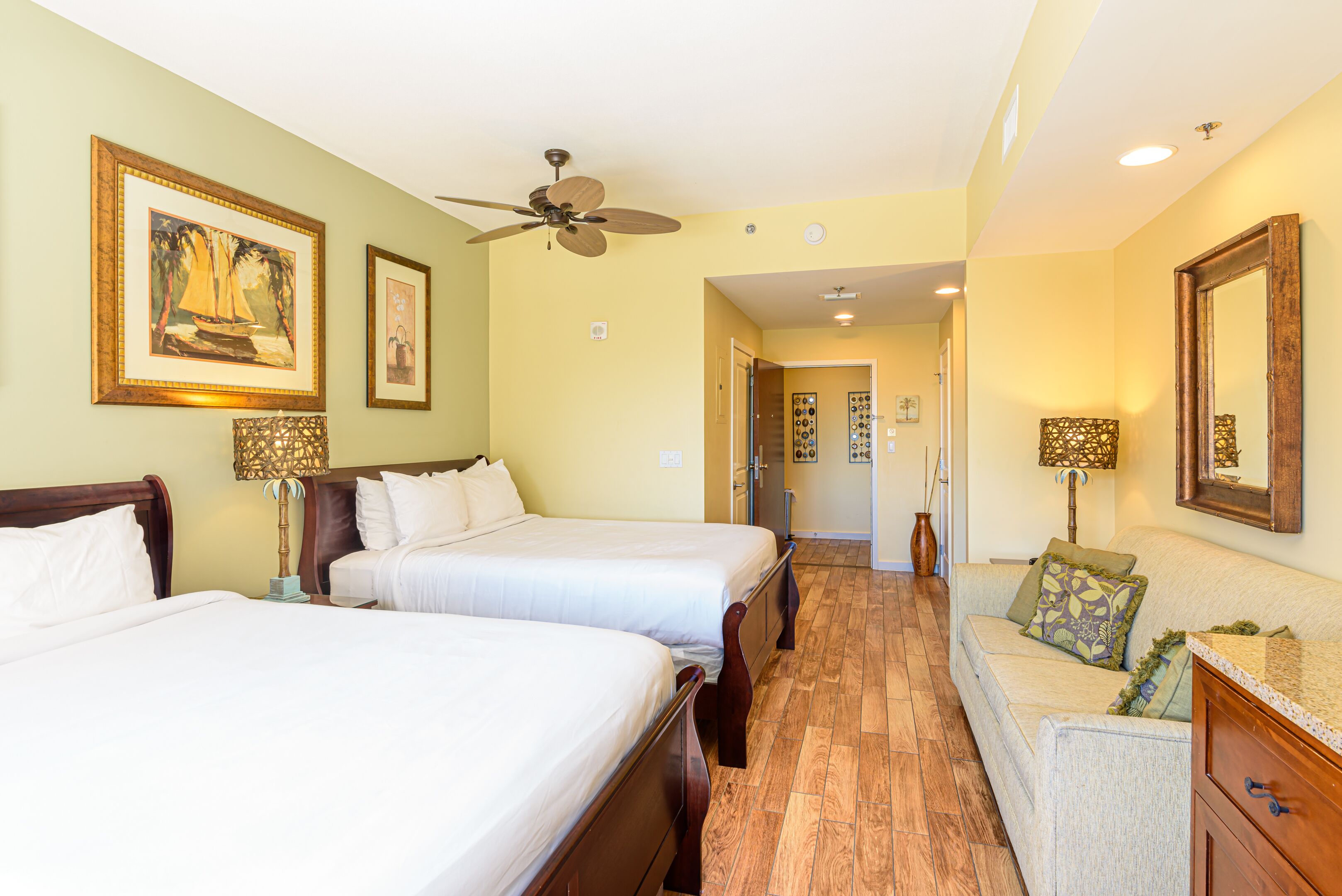 A hotel room with two beds, a sitting area, a ceiling fan, and artwork on the walls.