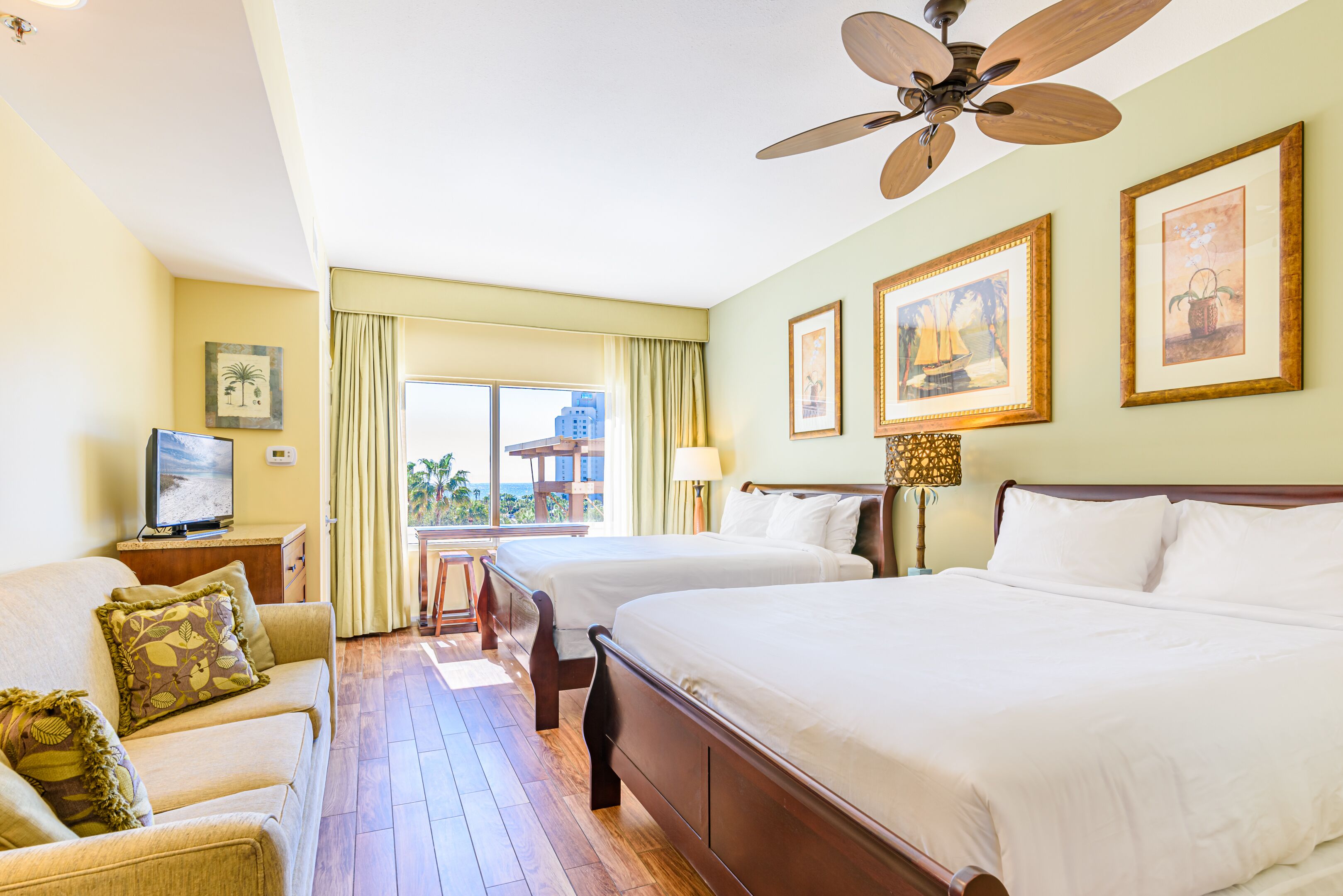 A hotel room with two beds, a sofa, a television, and a balcony with a view of the city.