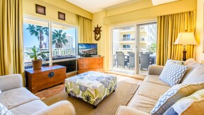 A hotel room with a sofa, a small table with a plant, a television, and a view of palm trees and a balcony. - Gulf Views ~ Private Balcony ~ Community Pool (Miramar Beach)