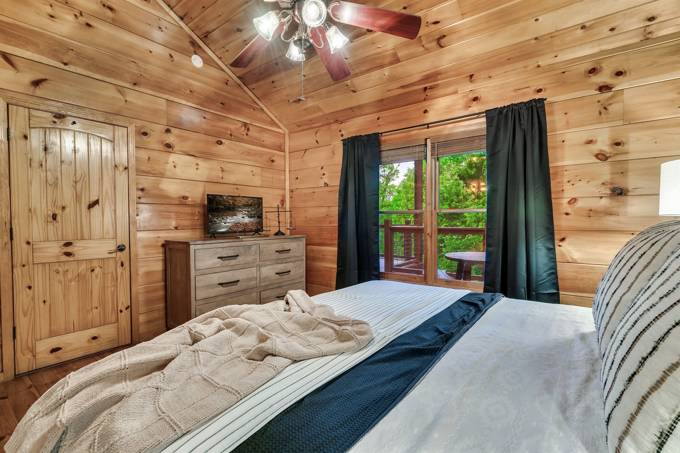 Ein Schlafzimmer in einer Holzhütte mit einem Bett, einem Schminktisch, einem Deckenventilator und einem Vorhangfenster.