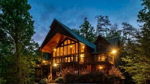 Une grande cabane en rondins à plusieurs étages, dotée d’un balcon imposant et de nombreuses fenêtres, et entourée d’une forêt dense.