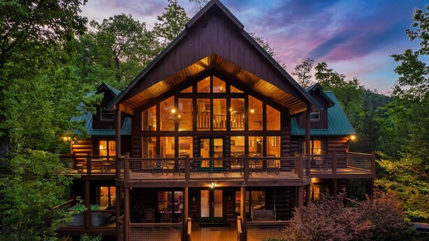 Une grande cabane en rondins à deux étages, dotée de nombreux balcons et de grandes fenêtres, et entourée d’une forêt dense.