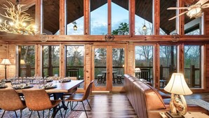 Un vaste espace dédié à la salle à manger, avec une longue table en bois, de nombreuses chaises et un banc. De grandes fenêtres offrent une vue sur l’extérieur.
